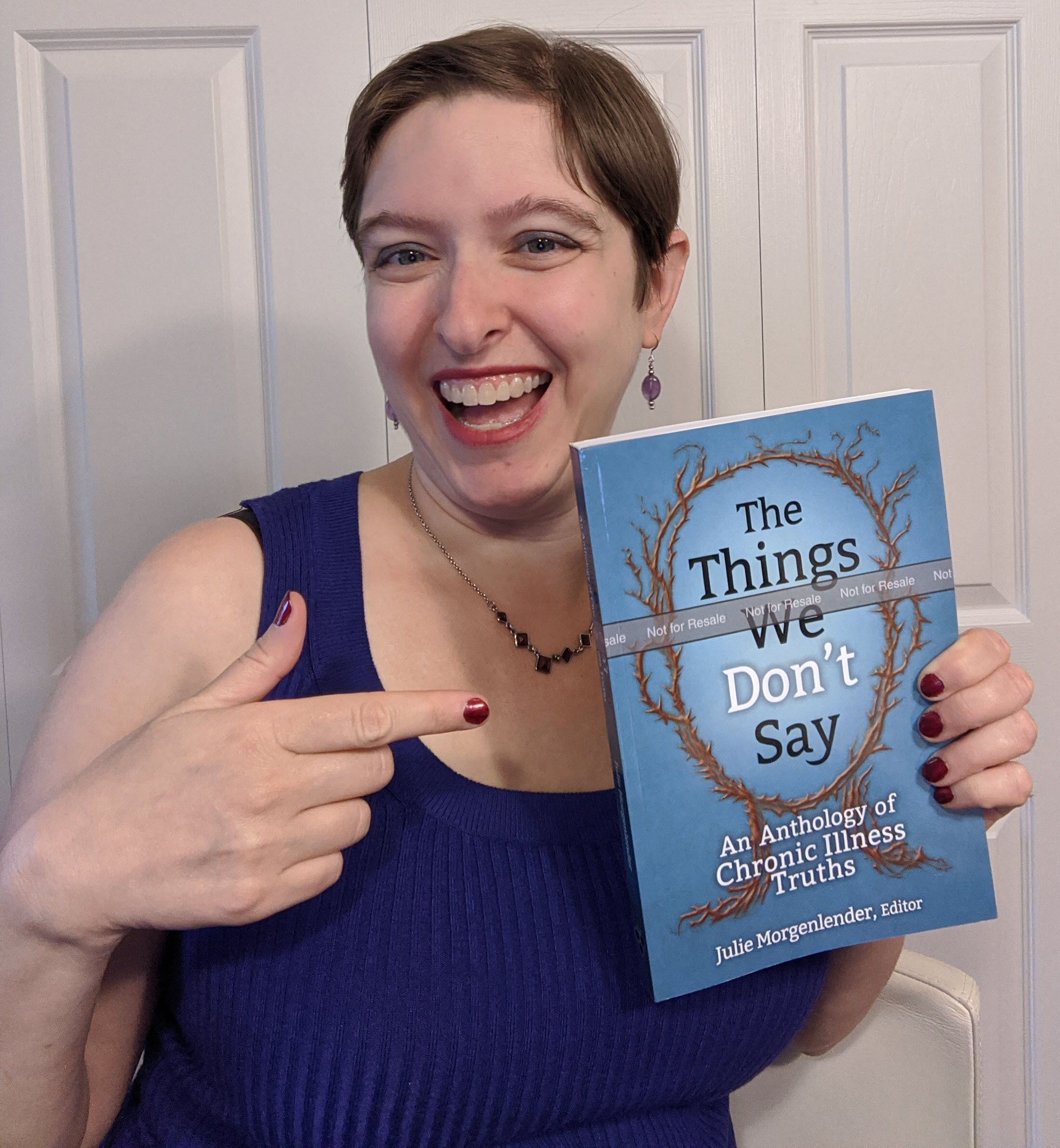 White woman with brown hair and blue shirt pointing with right hand at book in left hand with blue cover and title The Things We Don't Say: An Anthology of Chronic Illness Truths